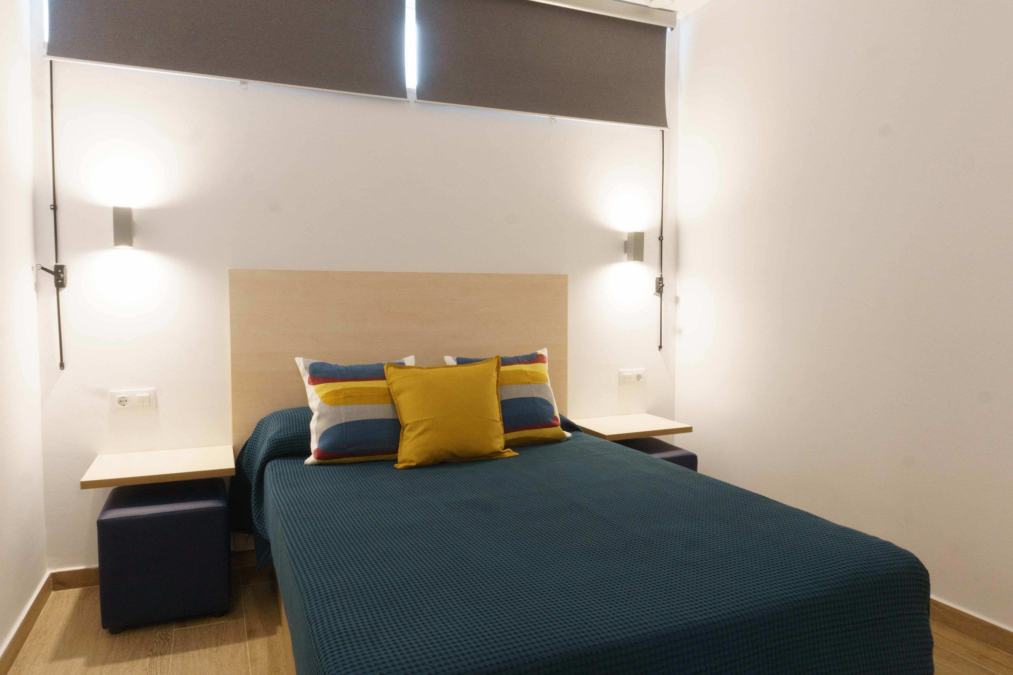 Una cama bien hecha con una colcha verde aguamarina y dos almohadas, una amarilla y otra multicolora, flanqueada por dos mesitas de noche con lámparas.