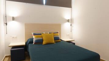 Una cama bien hecha con una colcha verde aguamarina y dos almohadas, una amarilla y otra multicolora, flanqueada por dos mesitas de noche con lámparas.