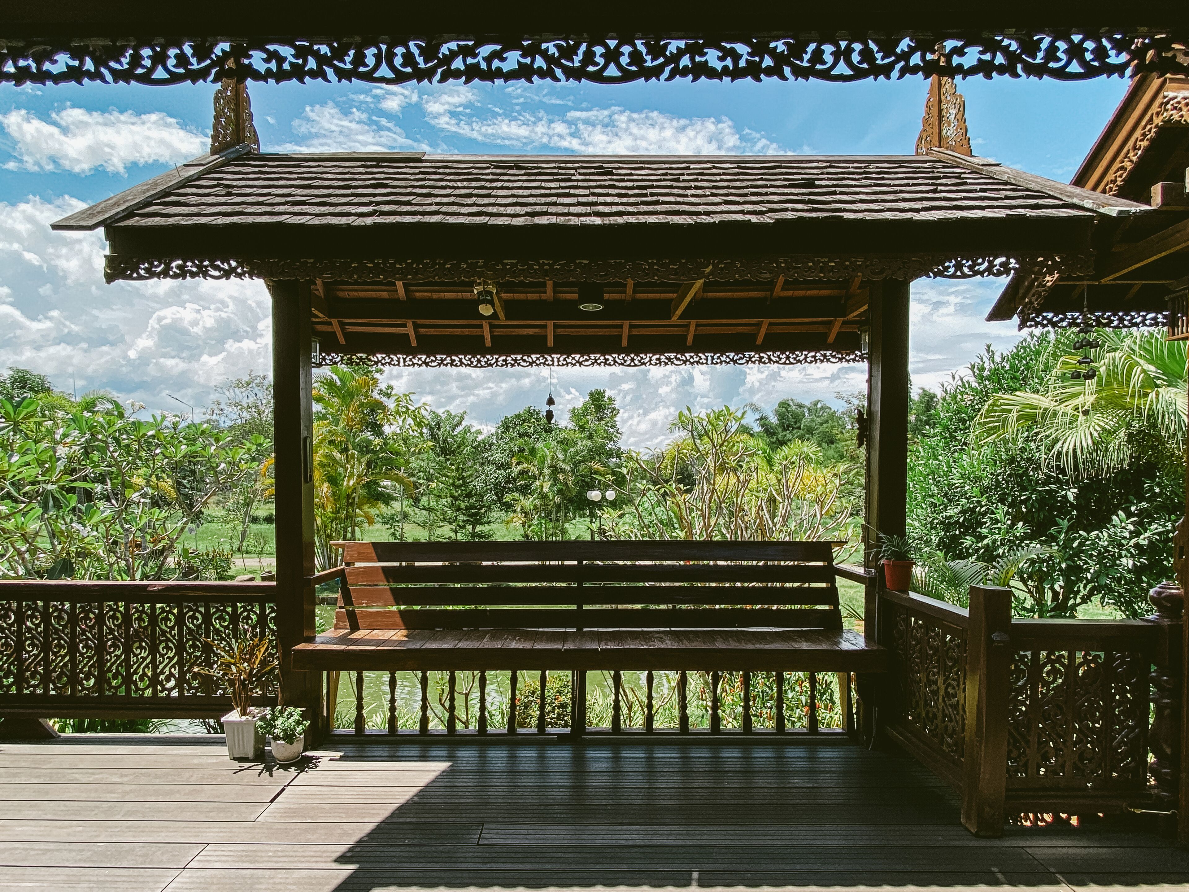 A wooden gazebo with a bench, surrounded by lush greenery and a clear sky.