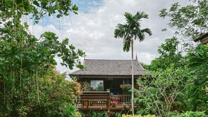 Una casa de madera con balcón, rodeada de abundante vegetación y un pequeño estanque.