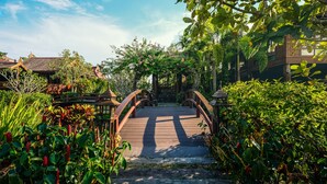 Un puente de madera que conduce a un edificio tradicional con techo de tejas, rodeado de abundante vegetación y plantas florecientes.