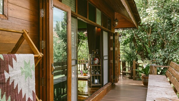 Una terraza de madera con un banco, una puerta de cristal que da a un cuarto con vistas a follaje, y un estante con cristalería.