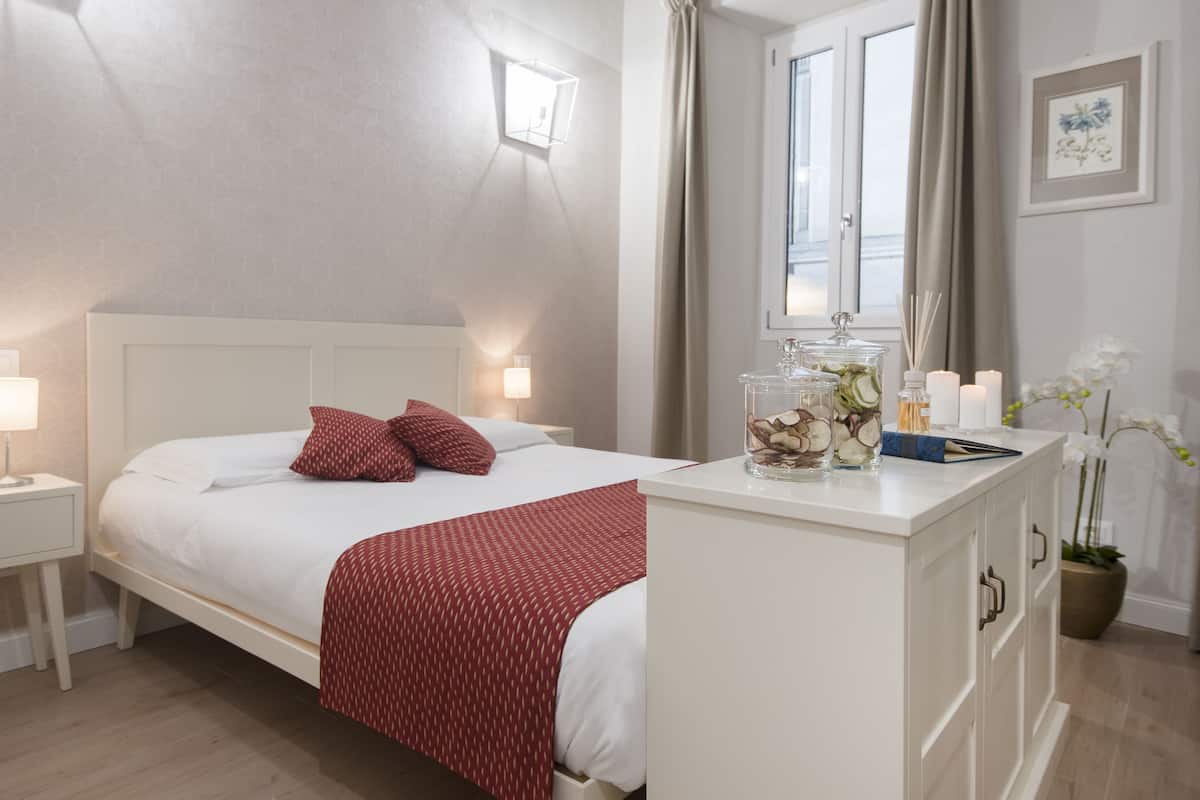 Een keurig opgemaakt slaapkamer met een bed, bedlampjes, een nachtkastje, een dressoir en een raam met gordijnen.