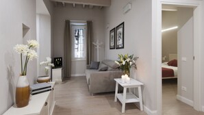A modern living room with a sofa, a small table with a candle, and a vase with flowers. There is a window with curtains and a coat rack in the background.