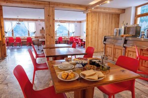 A dining area with wooden tables and red chairs, breakfast items on the table, and a kitchen area with a microwave and utensils. -  Miravalle Supersport Hotel (Forni Avoltri)