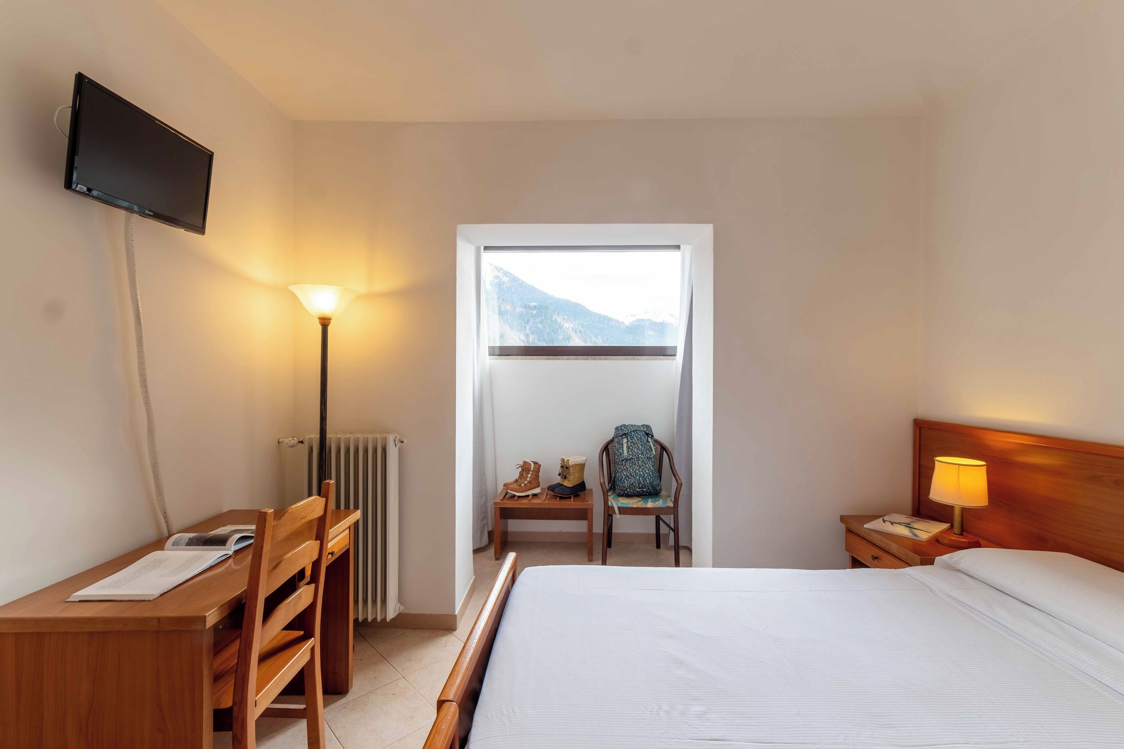 Habitación de hotel con cama, escritorio, silla, televisor, lámpara, mesita de noche y ventana con vista a la montaña.