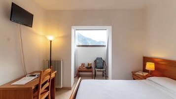 Habitación de hotel con cama, escritorio, silla, televisor, lámpara, mesita de noche y ventana con vista a la montaña.