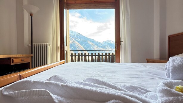 A hotel room with a bed, a view of mountains, a wooden headboard, and a radiator. -  Miravalle Supersport Hotel (Forni Avoltri)