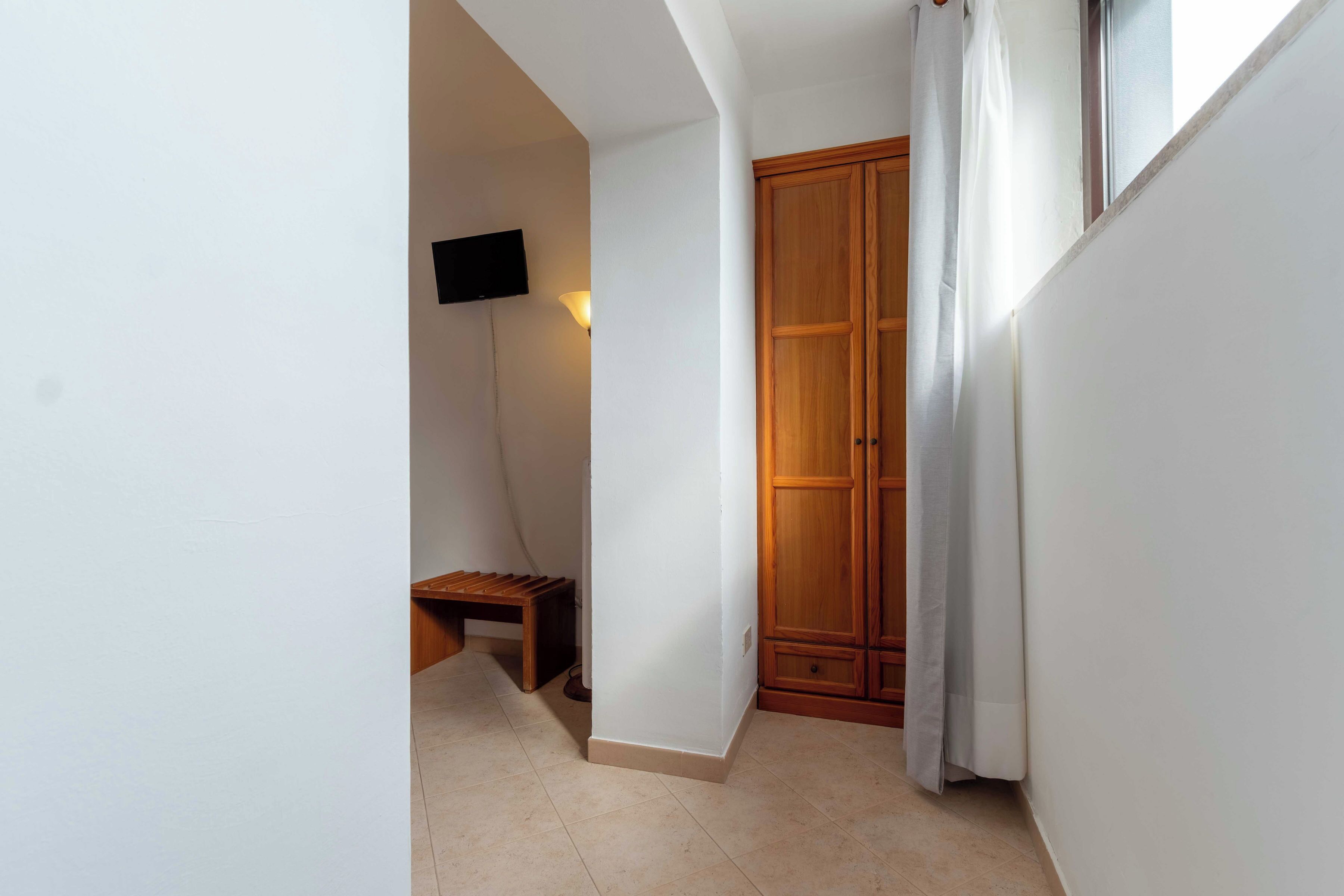 A narrow hallway with a wooden wardrobe, a bench, a wall-mounted TV, and a window with curtains.