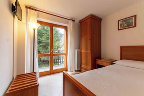 A bedroom with a bed, a wooden wardrobe, a nightstand, a lamp, a radiator, and a window with curtains. -  Miravalle Supersport Hotel (Forni Avoltri)