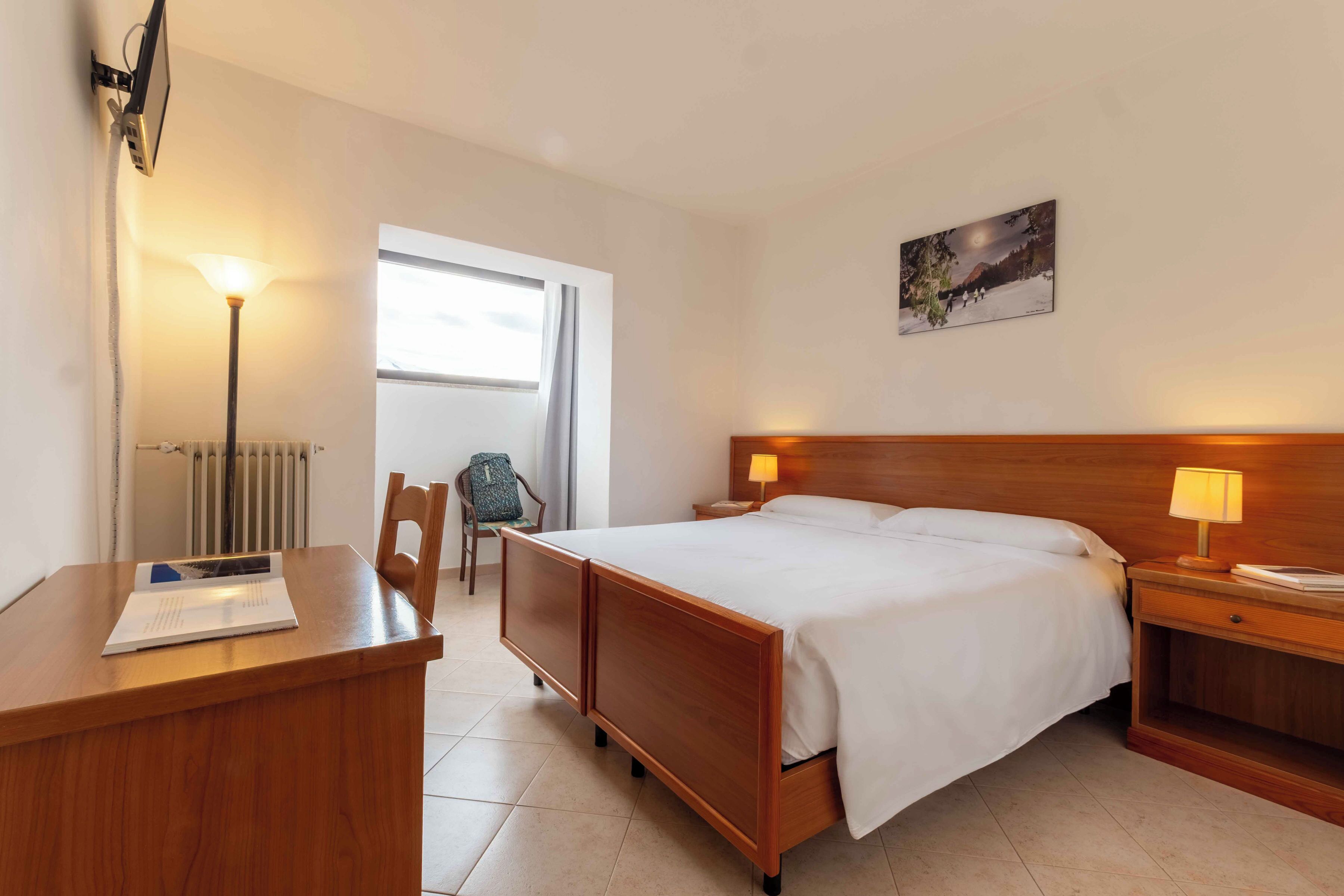 A hotel room with a bed, bedside lamps, a desk with a chair, a radiator, and a window.