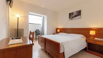 A hotel room with a bed, bedside lamps, a desk with a chair, a radiator, and a window.