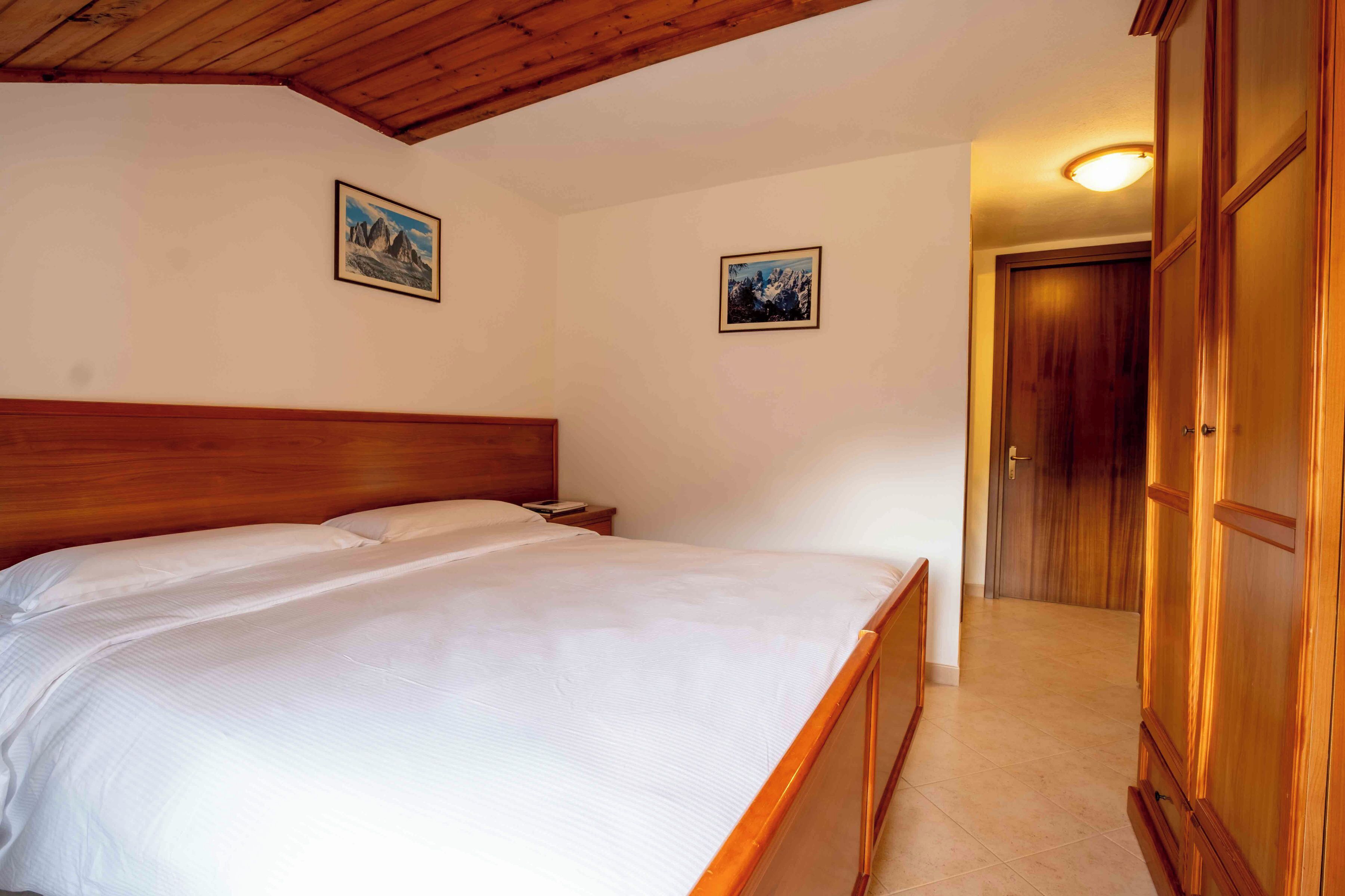 A bedroom with a wooden bed, a nightstand, two framed pictures on the wall, and a wooden wardrobe.