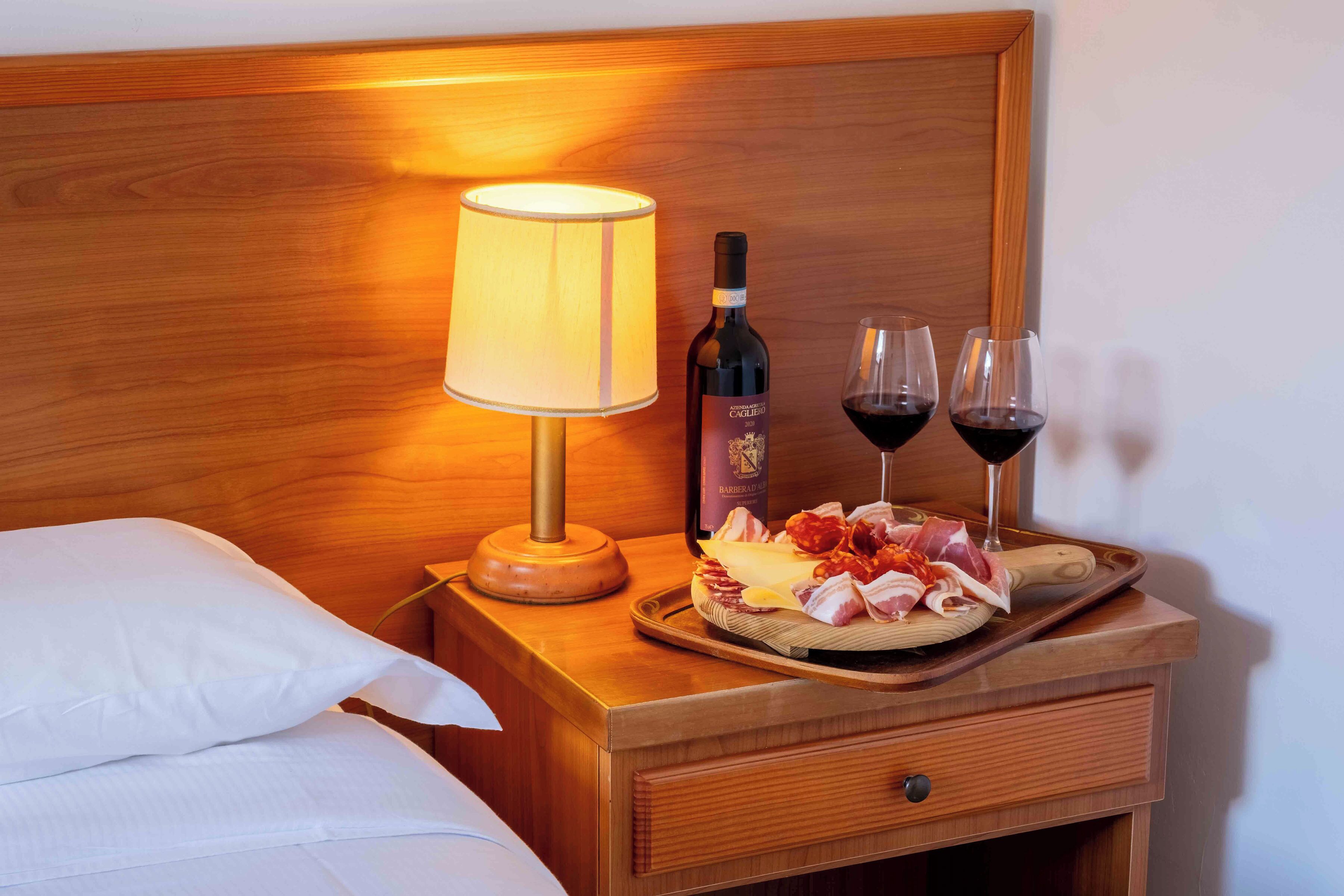 Een houten nachtkastje met een lamp, een fles wijn, glazen en een schaal vleeswaren.