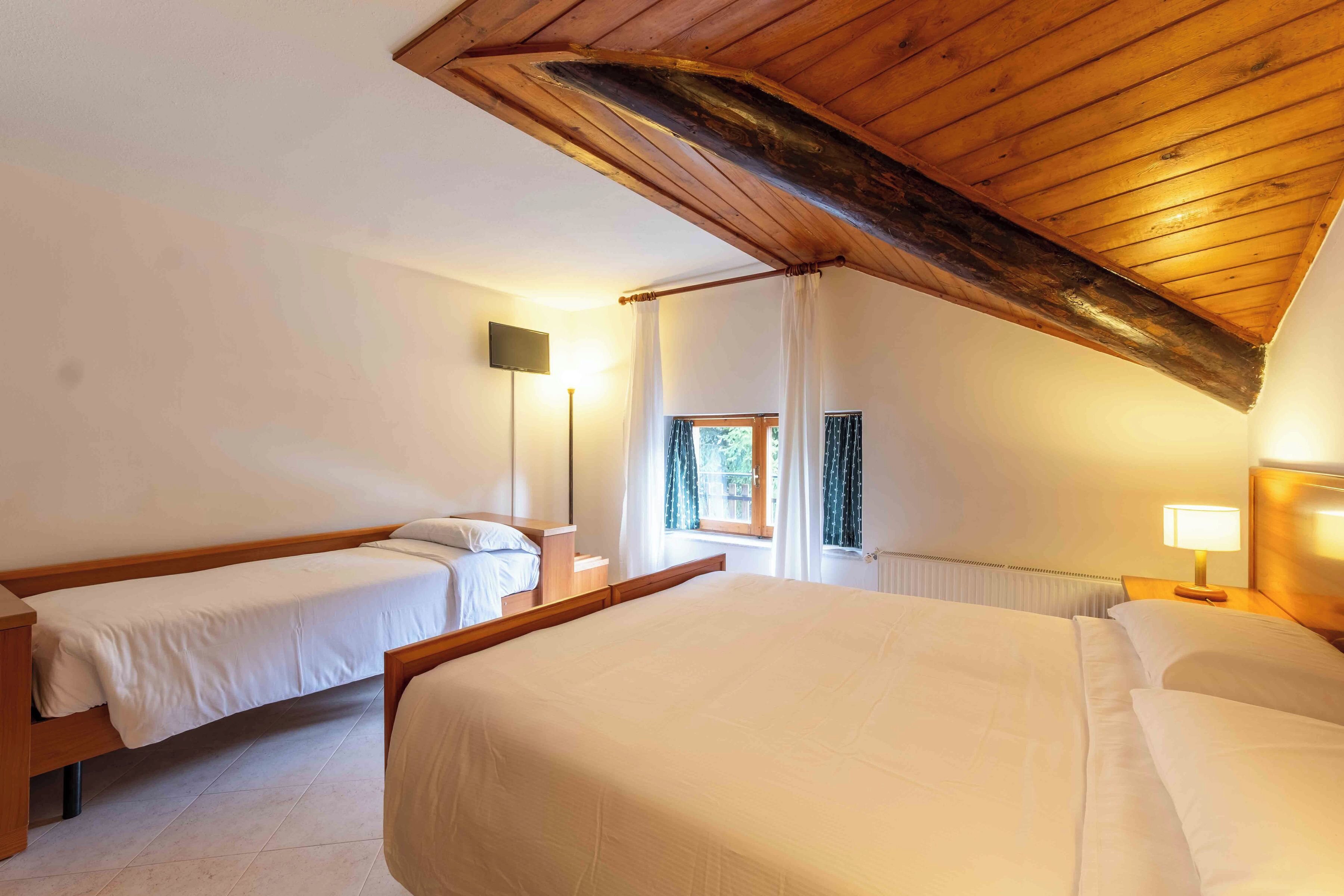 Un dormitorio con techo inclinado, dos camas individuales, una cama doble, una mesita de noche con lámpara, una ventana con cortinas y un televisor instalado en la pared.