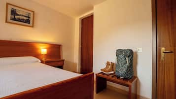 Habitación de hotel con cama de madera, lámpara de noche, un cuadro en la pared, un banco de madera y botas, y una puerta.