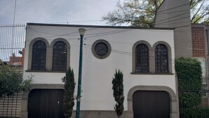 A building with three arched windows and a round window, a green street lamp, and a small tree in front. - Hotel Singular Coyoacán (Mexico City)