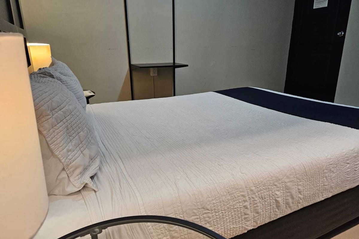 A single bed with a white quilt, a bedside lamp, and a small shelf.