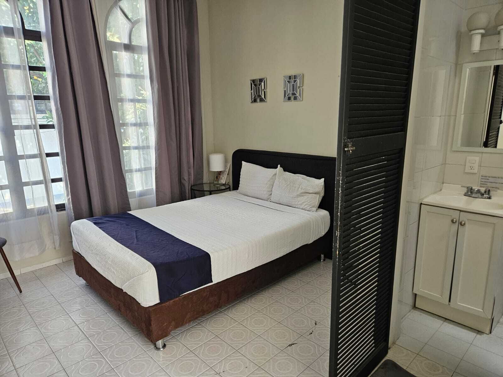 A bedroom with a bed, a nightstand, a lamp, a window with curtains, and a bathroom with a sink and cabinets.