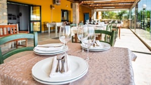 Mesa posta para dois, com pratos brancos, talheres e taças de vinho, em área de refeições ao ar livre, com paredes amarelas e cadeiras verdes.
