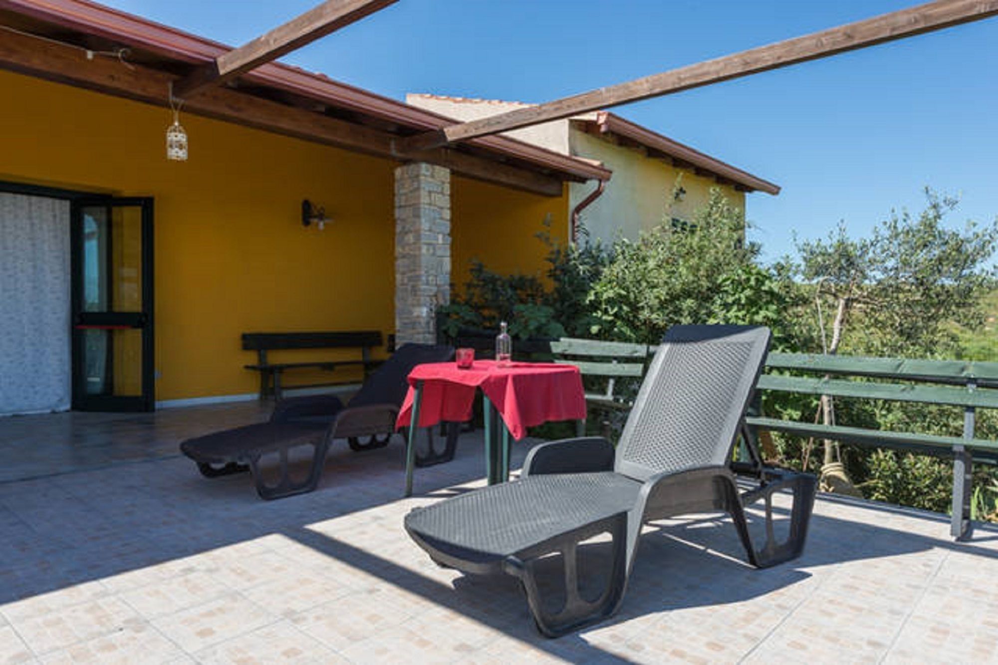 Una terrazza esterna con un edificio giallo, un pilastro in pietra, un tavolo rosso e due sedie.