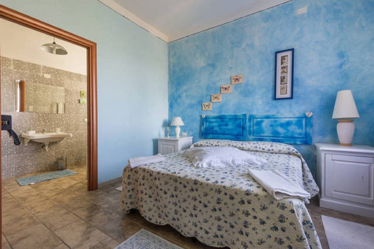 Ein Schlafzimmer mit einem Bett, einem Nachttisch, einer Lampe, einer Wand mit Bildern sowie einem Badezimmer mit Waschbecken und Dusche.