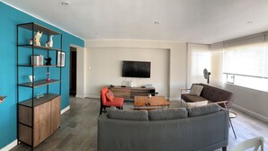 A modern living room with a grey sofa, a wooden coffee table, a red chair, and a teal bookshelf. A flat-screen TV is mounted on the wall, and there is a window providing natural light. - MLA apartments - 28 de Julio (Lima)