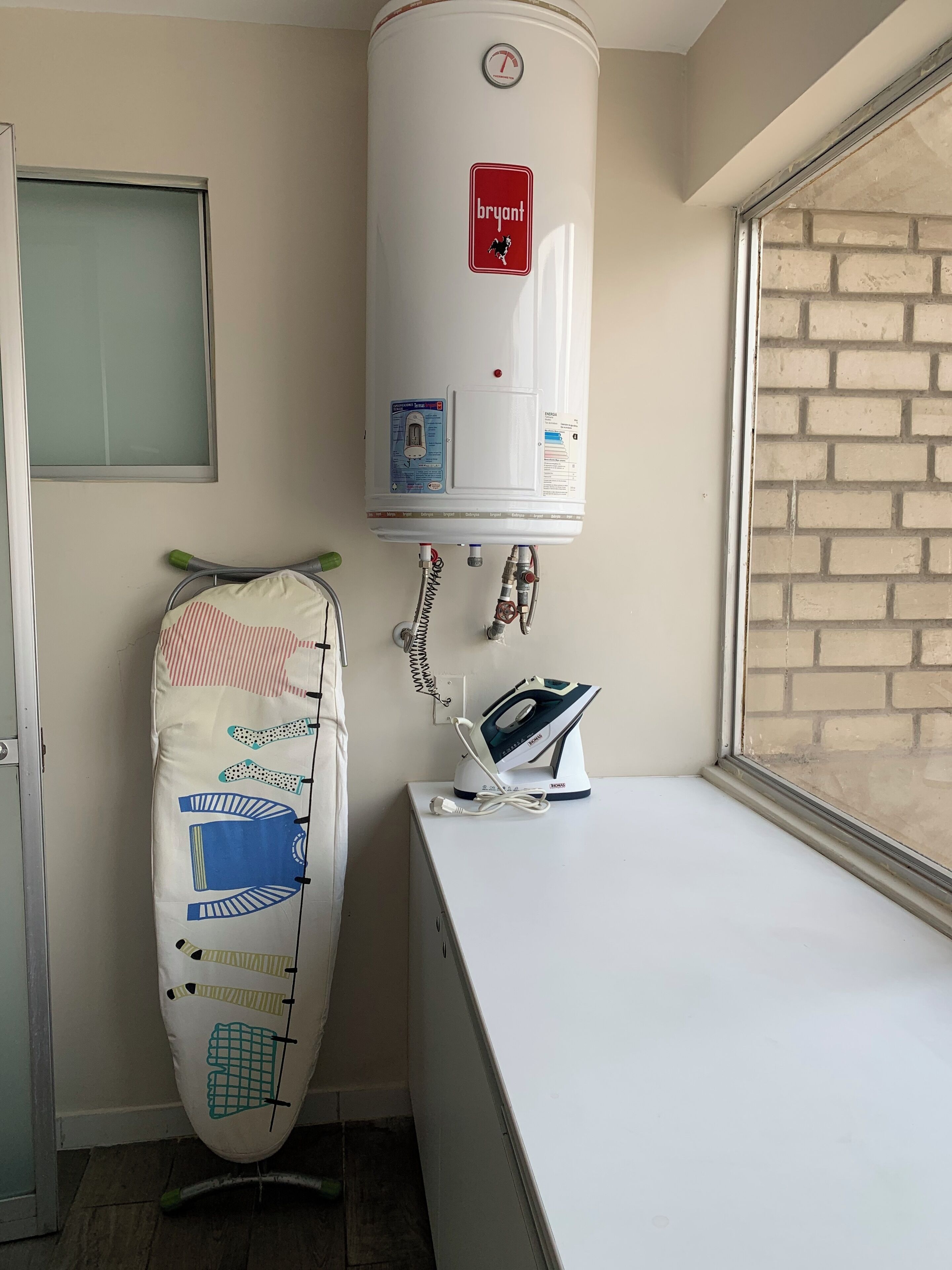 A compact room with a white radiator, a window with a brick wall view, and a small ironing station with an iron and a kettle.