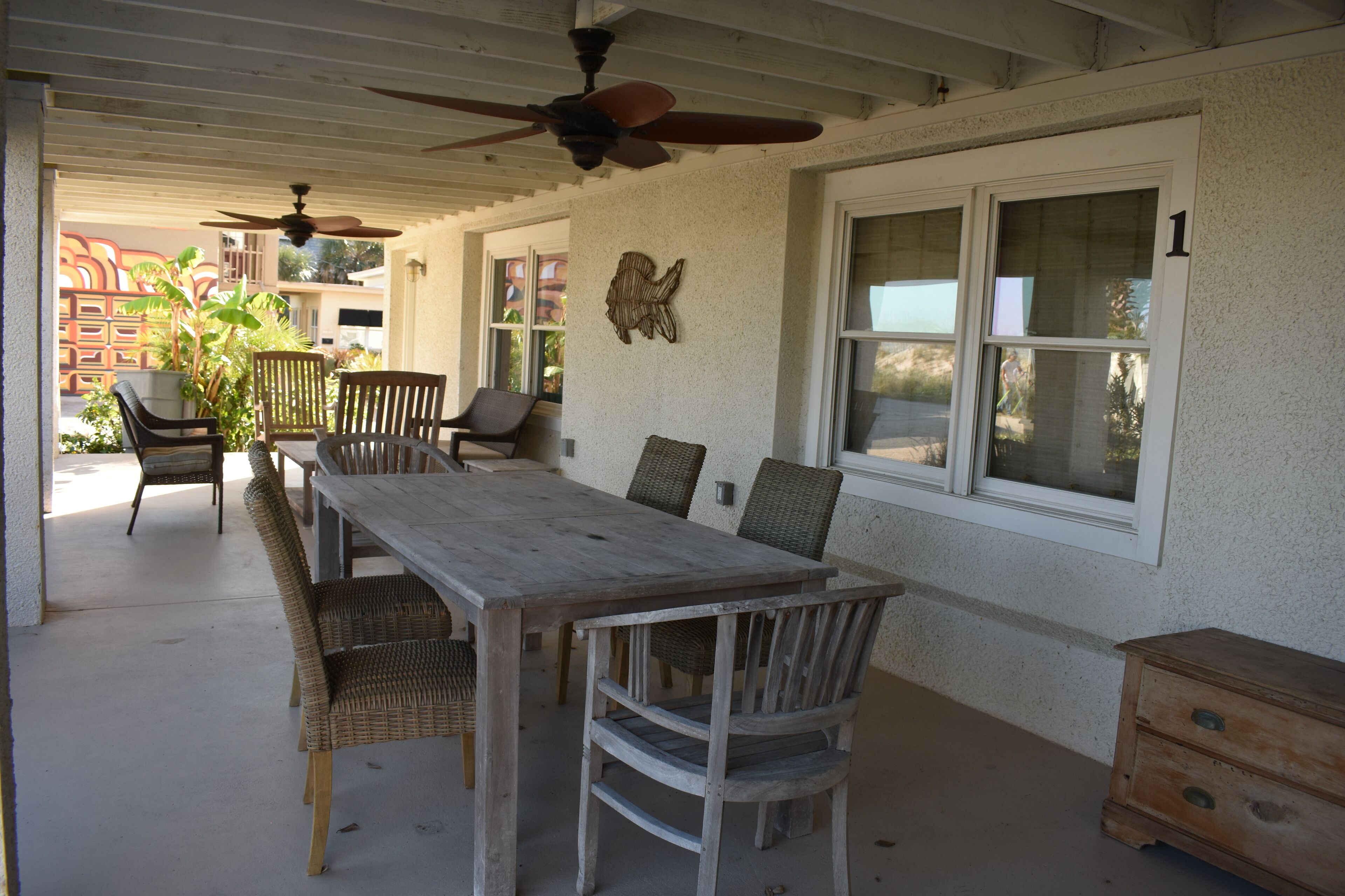 A covered patio with a wooden table and chairs, a ceiling fan, and a decorative wall piece.