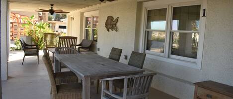 A covered patio with a wooden table and chairs, a ceiling fan, and a decorative wall piece.