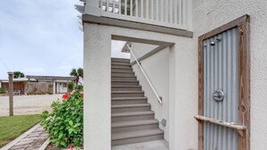 Una escalera de concreto lleva a una puerta de madera con manija de metal, rodeada por un muro blanco y un pequeño jardín con flores rojas.