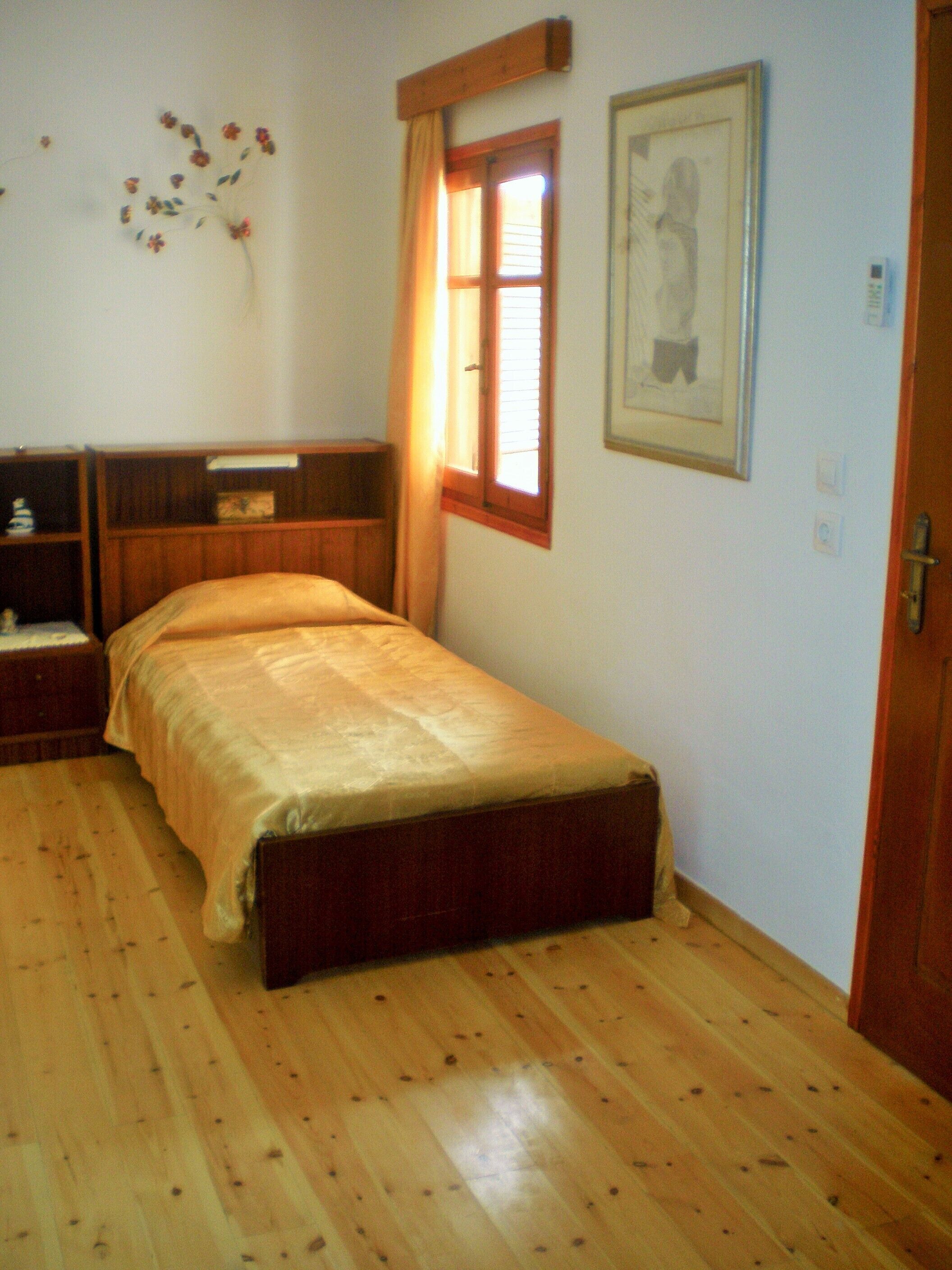 Une petite chambre à coucher, bien rangée, avec un lit simple, une table de chevet, une armoire en bois, une fenêtre avec des rideaux et un tableau encadré accroché au mur.