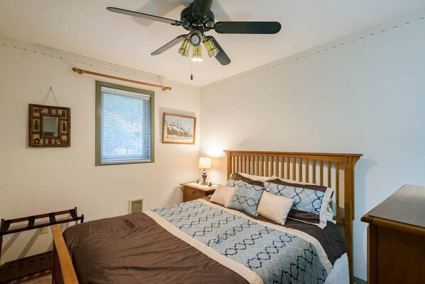 Una camera da letto con un letto, un comodino, una lampada da comodino, una ventola a soffitto, una finestra con persiane e un quadro appeso al muro.