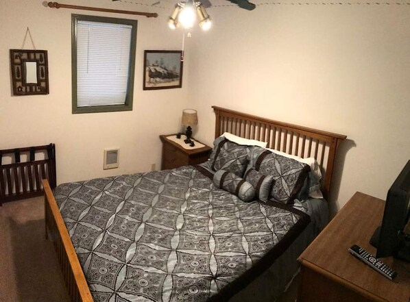 Une chambre à coucher comprenant un lit, une table de chevet, une chaise, une fenêtre avec des stores et un ventilateur de plafond.