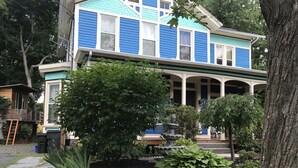 Una casa de dos pisos con fachada azul y blanca, un porche cubierto y un jardín con diversas plantas y una fuente.