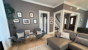 Una sala moderna con un sofá gris, dos sillones, una mesa de centro y una alfombra estampada. Hay cuadros enmarcados en la pared y una escalera que lleva a otra habitación.