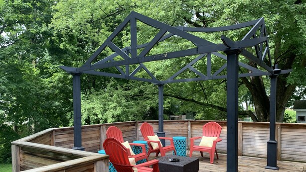 Una terraza de madera con sillas Adirondack rojas y una mesa, rodeada por una pérgola de metal.