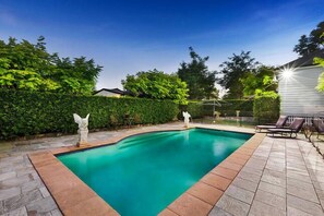A rectangular swimming pool with a stone border, surrounded by a patio area with lounge chairs and a hedge.