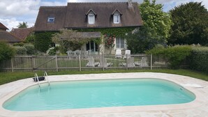 Uma piscina com um deck de pedra, rodeada por uma cerca de madeira, e uma casa com telhado inclinado e janelas ao fundo.
