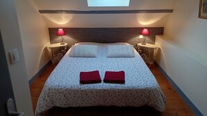 Quarto com claraboia, duas mesas de cabeceira com luminárias, cama com colcha floral e duas toalhas vermelhas, além de piso de madeira.