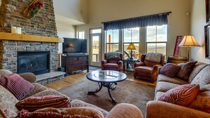 A spacious living room with a stone fireplace, leather sofas, a glass coffee table, and large windows offering a view of the outdoors. - 1 Mi to Granby Ranch: Mtn Home w/ Deck (Granby)