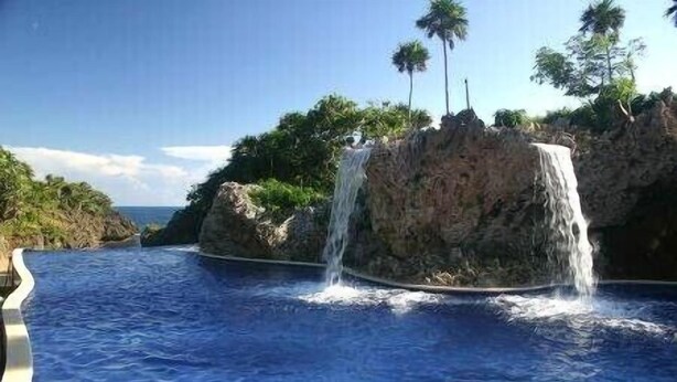 A pool with a waterfall feature surrounded by tropical vegetation and a clear sky. - Luxury 2 Bedroom, 2.5 Bath Dream Rental In West Bay (West Bay)