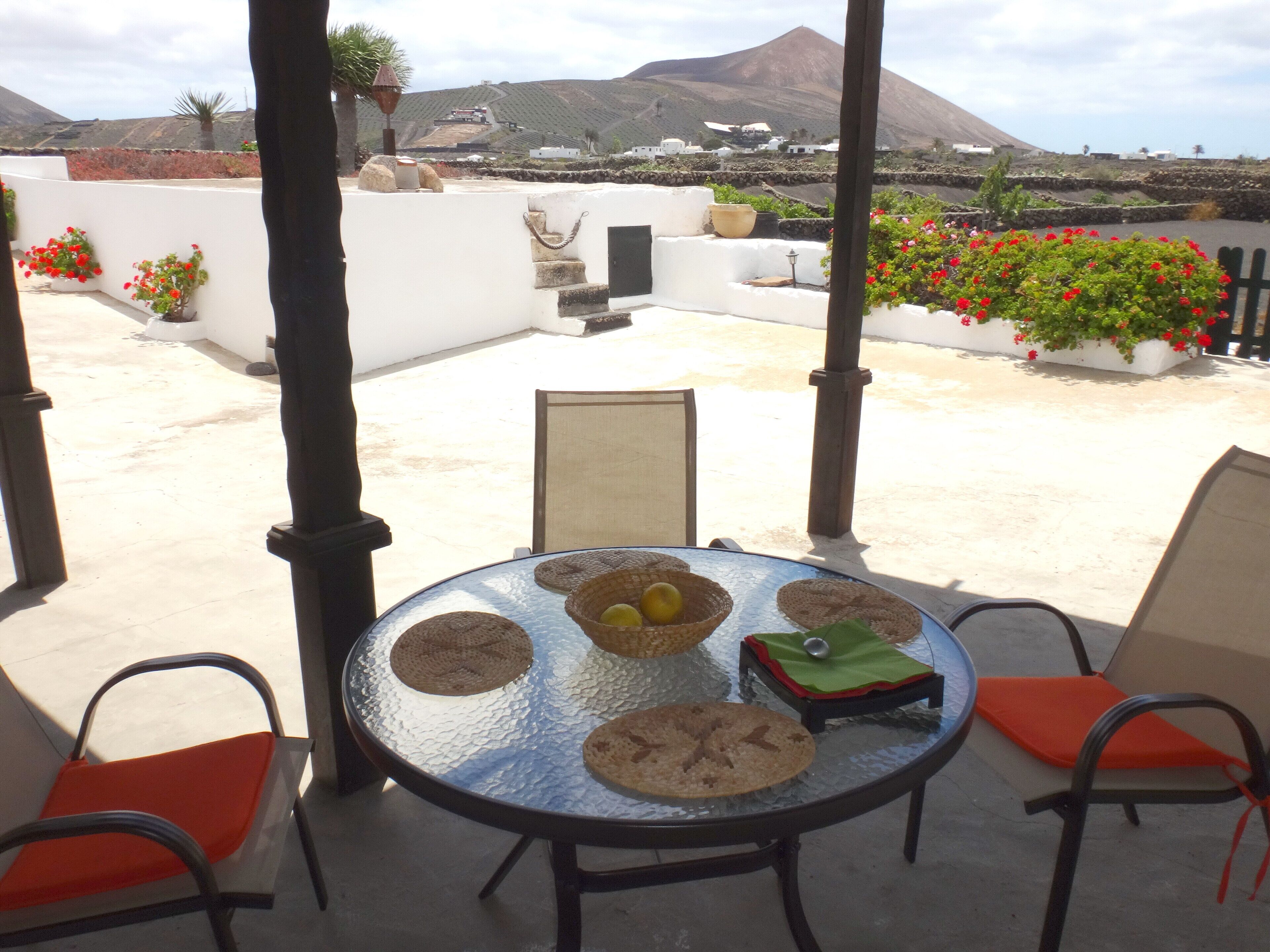 Een ronde tafel met vier geweven placemats, twee stoelen met oranje kussens, en een bergachtig landschap op de achtergrond.