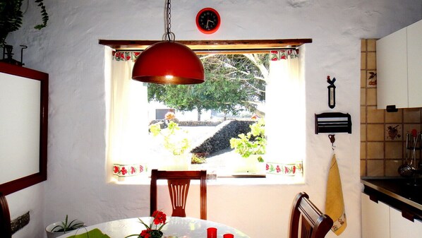 A dining area with a table, chairs, a hanging red lamp, a window with a view of greenery, and a wall clock. - In the Heart of Lanzarote! (Masdache)