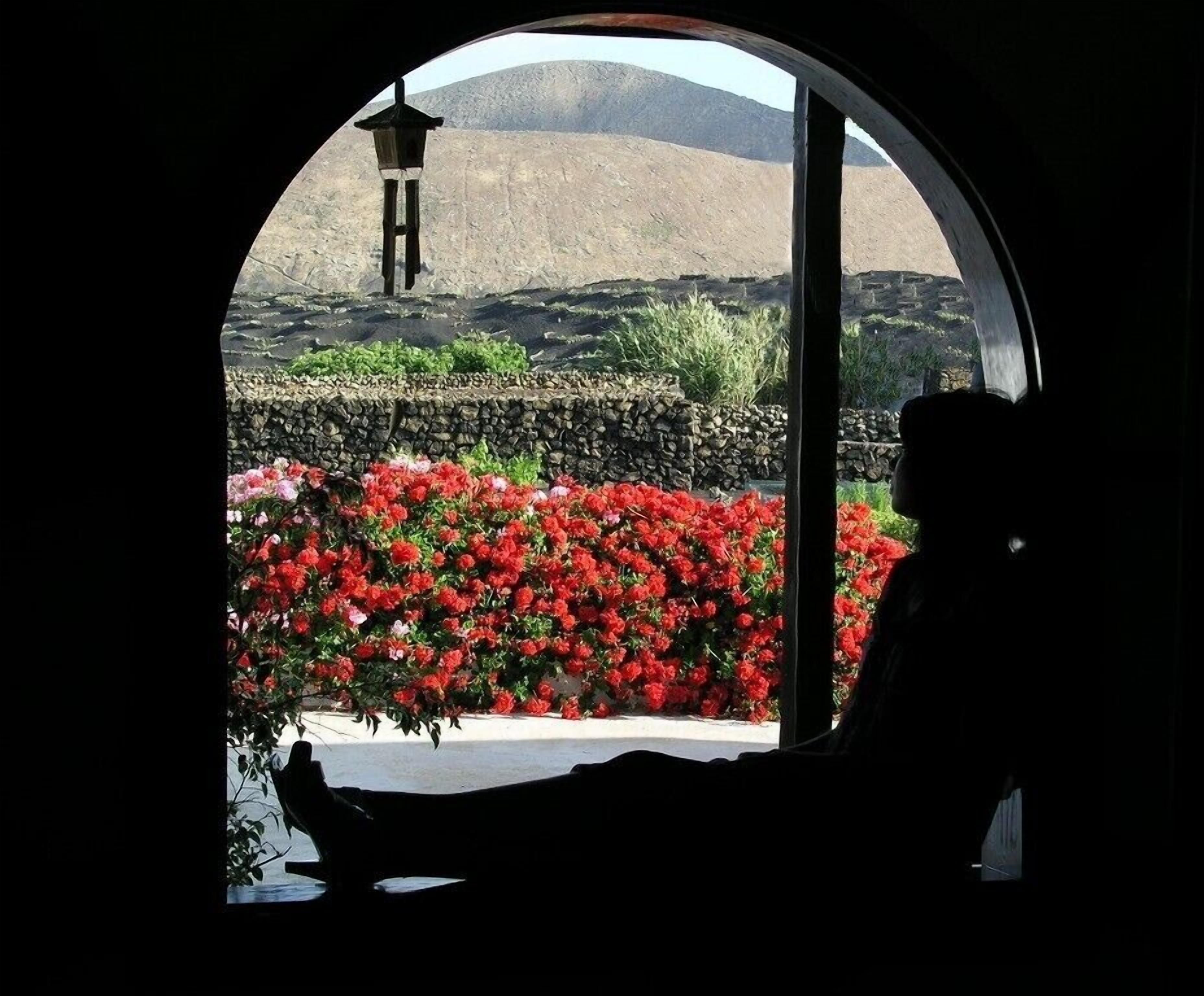 A view through a circular window of a stone wall, a red flower bed, and a silhouette of a person.