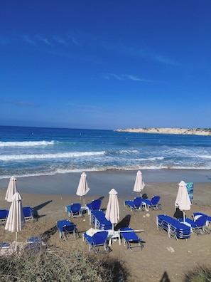 Una playa con hileras de sillas azules y sombrillas blancas, una orilla arenosa y el océano al fondo.