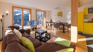 A modern living room with a brown sofa, a dining area with a wooden table and chairs, and a kitchen with yellow cabinets. - Chalet Wyssbirg (Wengen)