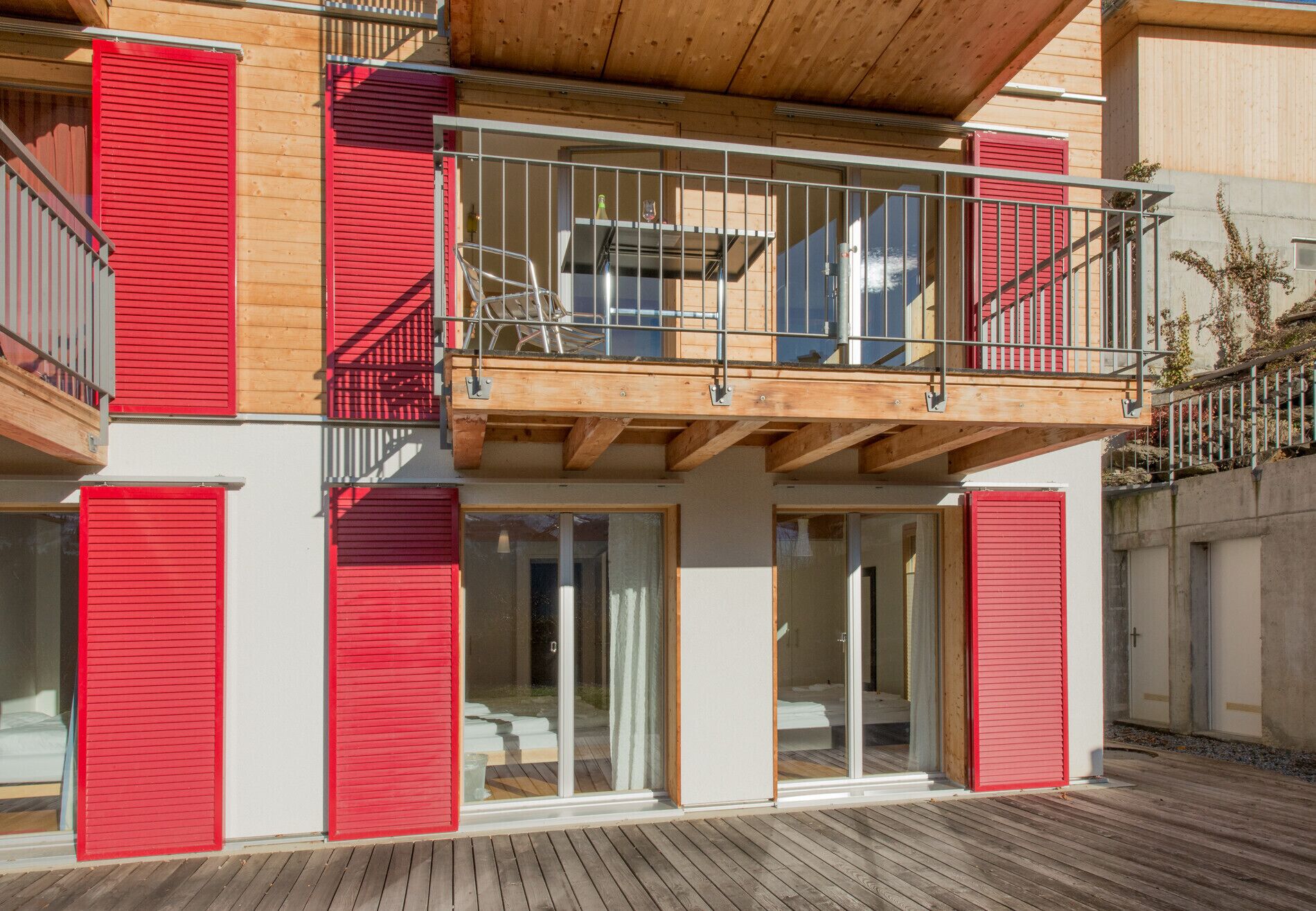 Un bâtiment moderne à deux étages, doté de volets rouges, d’un balcon avec des balustrades en métal et d’une terrasse en bois.