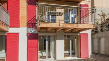 Un bâtiment moderne à deux étages, doté de volets rouges, d’un balcon avec des balustrades en métal et d’une terrasse en bois.
