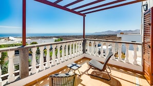 Un balcón con barandilla blanca, muebles de mimbre y vistas al mar y a las montañas.